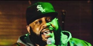 A man in a hoodie, jacket and baseball cap speaks into a microphone