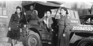 four black women in army uniforms stand in front of a truck
