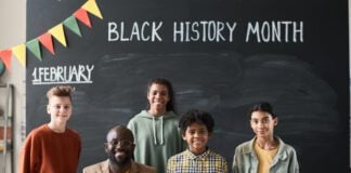 Portrait of African American teacher smiling at camera together with his children while they standing against the blackboard